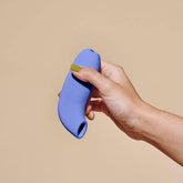 A hand with painted nails holds a blue, curved, cylindrical object with a small opening at the end—a Dame Aer - Air Pulse Clit Stimulator by Dame—against a beige background.